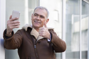 Ceketli olgun bir adam telefonla selfie çekiyor ve parmağıyla bir sorun yaratmıyor.