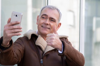 Ceketli olgun bir adam telefonla selfie çekiyor ve parmağıyla bir sorun yaratmıyor.