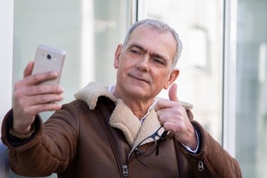 Ceketli olgun bir adam telefonla selfie çekiyor ve parmağıyla bir sorun yaratmıyor.