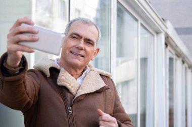Kışlık ceketli olgun bir adam telefonla selfie çekiyor.