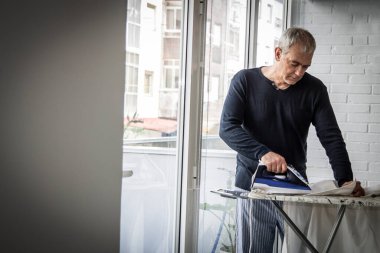 Evde ütü yapan olgun bir adam, ev işlerinde konsept eşitliği sağlar.