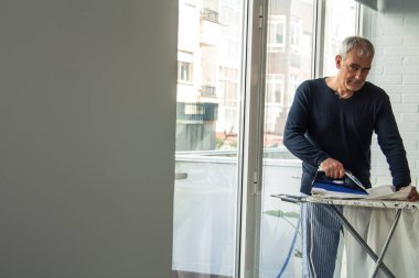 Evde ütü yapan olgun bir adam, ev işlerinde konsept eşitliği sağlar.