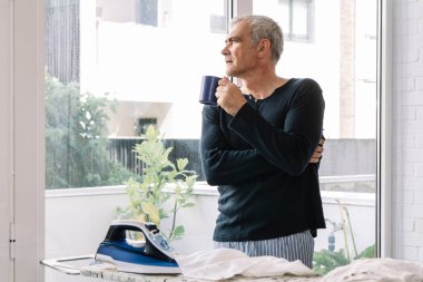 Adam evde gömleğini ütülerken kahve içiyor.