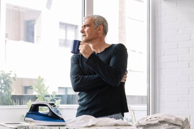 Adam evde gömleğini ütülerken kahve içiyor.