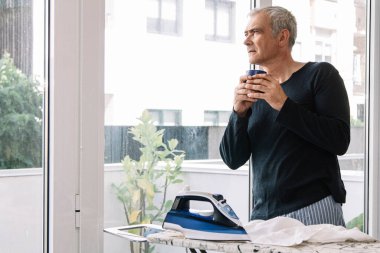 Adam evde gömleğini ütülerken kahve içiyor.