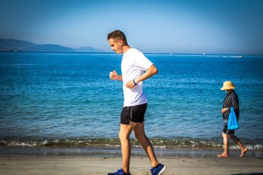 Genç bir adam önlem olarak arka planda tıbbi maske takan bir kadınla plaj boyunca koşuyor.