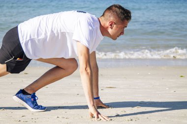 Genç adam sahilde koşmak için başlangıç pozisyonunda.