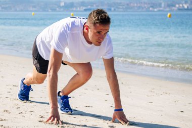 Genç adam sahilde koşmak için başlangıç pozisyonunda.