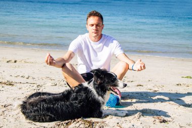 Plajda köpeğiyle yoga yapan bir adam.