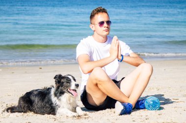 Plajda köpeğiyle yoga yapan bir adam.