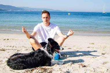 Plajda köpeğiyle yoga yapan bir adam.