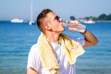 Genç adam plajda dinleniyor ve egzersiz yaptıktan sonra içiyor.