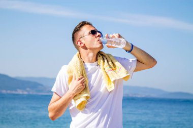 Genç adam plajda dinleniyor ve egzersiz yaptıktan sonra içiyor.