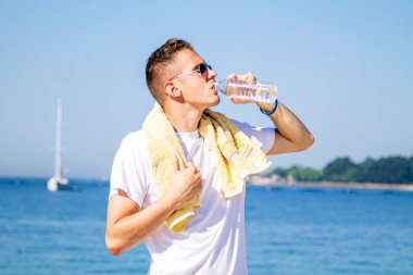 Genç adam plajda dinleniyor ve egzersiz yaptıktan sonra içiyor.