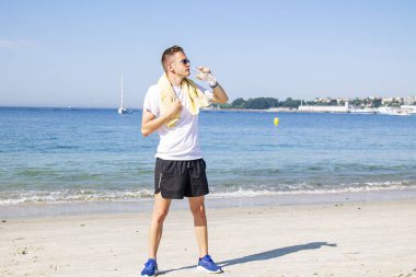 Genç adam plajda dinleniyor ve egzersiz yaptıktan sonra içiyor.