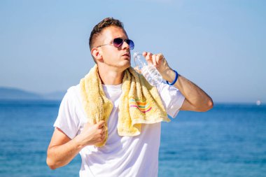 Yorgun adam su içiyor ve spordan sonra sahilde dinleniyor.