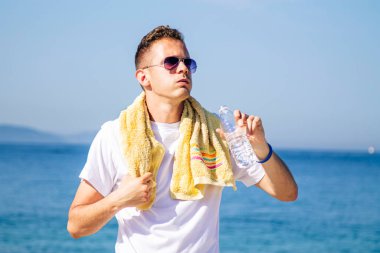 Yorgun adam su içiyor ve spordan sonra sahilde dinleniyor.