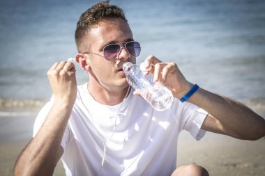 Sahilde dinlenen bir adam spor yaptıktan sonra kulaklıkla ve cep telefonuyla müzik dinliyor.