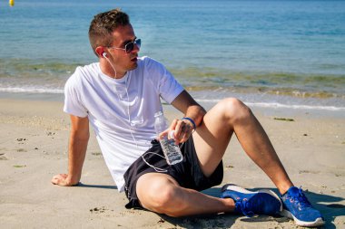 Sahilde dinlenen bir adam spor yaptıktan sonra kulaklıkla ve cep telefonuyla müzik dinliyor.