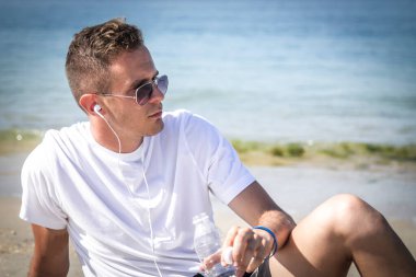 Adam plajda dinleniyor. Koşudan sonra içerken kulaklıkla ve cep telefonuyla müzik dinliyor.