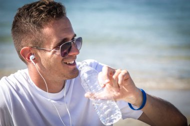 Adam plajda dinleniyor. Koşudan sonra içerken kulaklıkla ve cep telefonuyla müzik dinliyor.