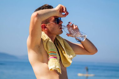 Adam egzersiz yaptıktan sonra sahilde dinleniyor ve su içiyor.