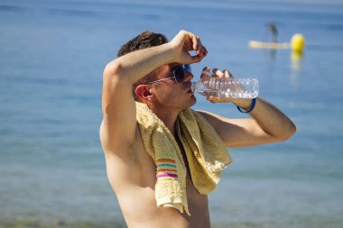 Adam egzersiz yaptıktan sonra sahilde dinleniyor ve su içiyor.