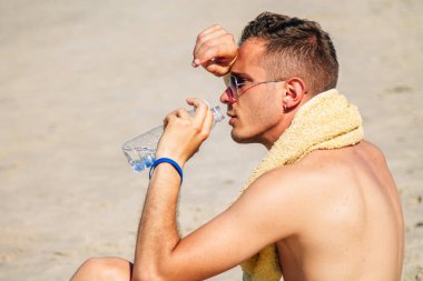 Adam egzersiz yaptıktan sonra sahilde dinleniyor ve su içiyor.