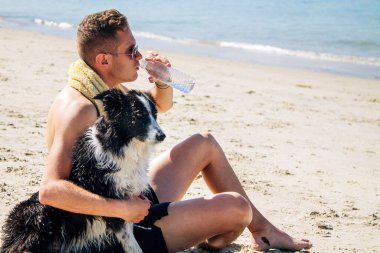 Genç adam sahilde köpeğiyle dinleniyor ve içiyor.
