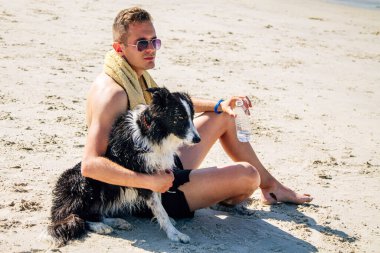 Genç adam sahilde köpeğiyle dinleniyor ve içiyor.