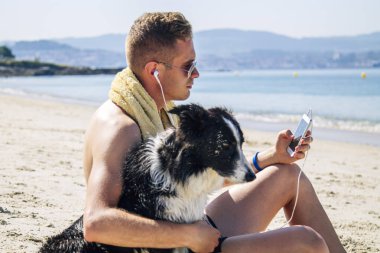 Kumsalda evcil hayvanıyla rahat bir adam cep telefonu kulaklıklarıyla müzik dinliyor.