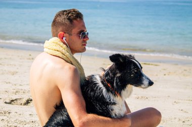 Genç bir adam köpeğiyle sahilde kulaklıkla müzik dinliyor.