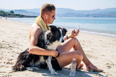 Kumsalda evcil hayvanıyla rahat bir adam cep telefonu kulaklıklarıyla müzik dinliyor.