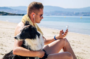Kumsalda evcil hayvanıyla rahat bir adam cep telefonu kulaklıklarıyla müzik dinliyor.