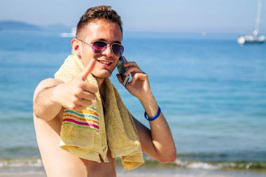 Genç adam sahilde cep telefonuyla seyahat ve otel rezervasyonu yapıyor.