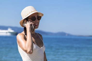 Plajda şapkalı bir kadın cep telefonuyla konuşuyor.
