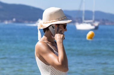 Plajda şapkalı bir kadın cep telefonuyla konuşuyor.