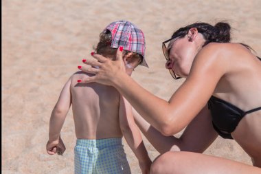 Anne plajdaki çocuğa güneş kremi sürüyor.