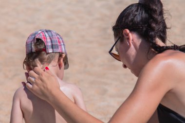 Anne plajdaki çocuğa güneş kremi sürüyor.