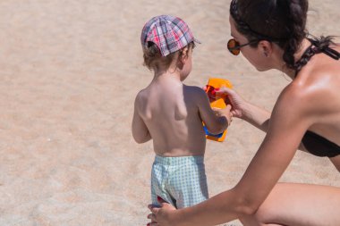 Anne plajdaki çocuğa güneş kremi sürüyor.