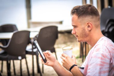 Adam dijital tablet ya da kitap kullanıyor ve dışarıda kahve içiyor.