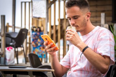 Genç adam cep telefonu kullanıyor ve açık kafede kahve içiyor.