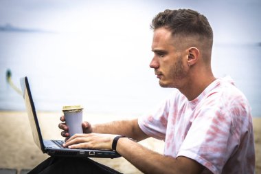 Genç adam dışarıda bilgisayarını kullanırken kahve içiyor.