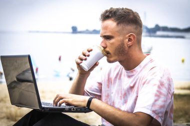 Genç adam dışarıda bilgisayarını kullanırken kahve içiyor.