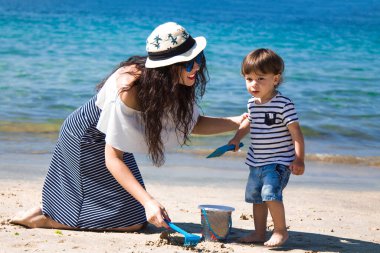 Anne ve küçük oğlu sahilde kova ve kürekle oynuyorlar.