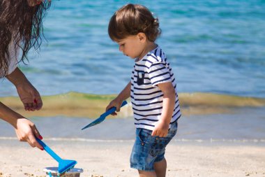 Anne ve küçük oğlu sahilde kova ve kürekle oynuyorlar.