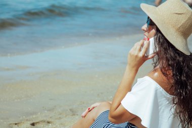 Şapkalı bir kadın kumsalda cep telefonuyla konuşuyor.