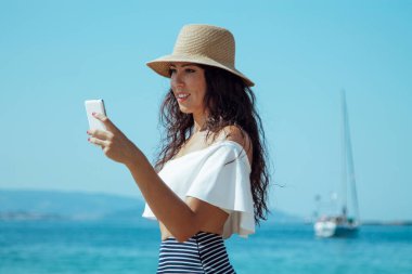 Plajdaki şapkalı kadın cep telefonunu kontrol ediyor.