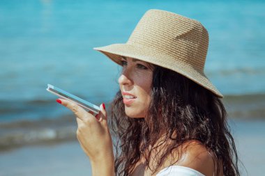 Plajda şapkalı bir kadın cep telefonuyla mesaj yolluyor.