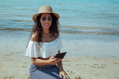 Sahilde şapkalı genç bir kadın dijital tablet ya da e-kitap kullanıyor.
